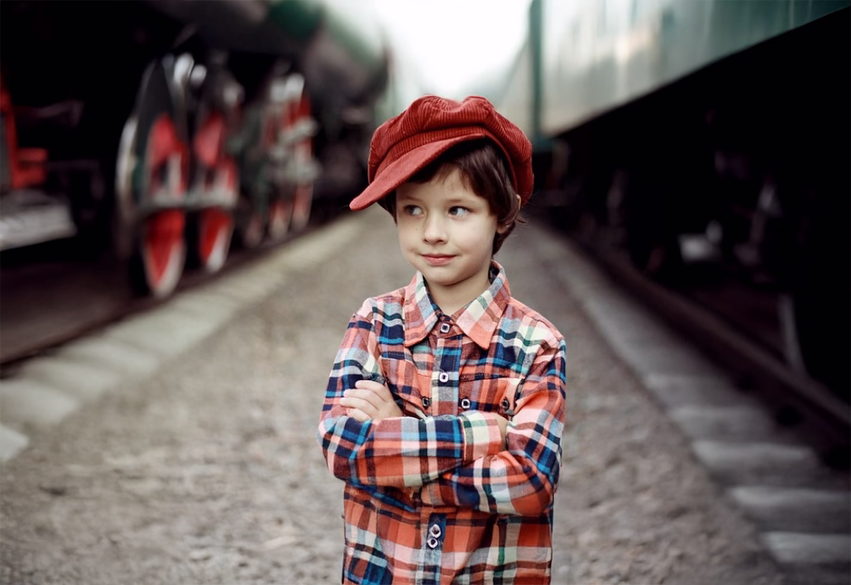 Chłopiec w czerwonej czapce i kolorowej koszuli w kratę stoi przy torach kolejowych. Z pewnym siebie wyrazem twarzy i skrzyżowanymi ramionami spogląda w bok. W tle widoczny fragment pociągu. Zdjęcie symbolizuje ciekawość poznawczą i odwagę, cechy rozwijane podczas warsztatów dla uczniów w Poradni Expert.